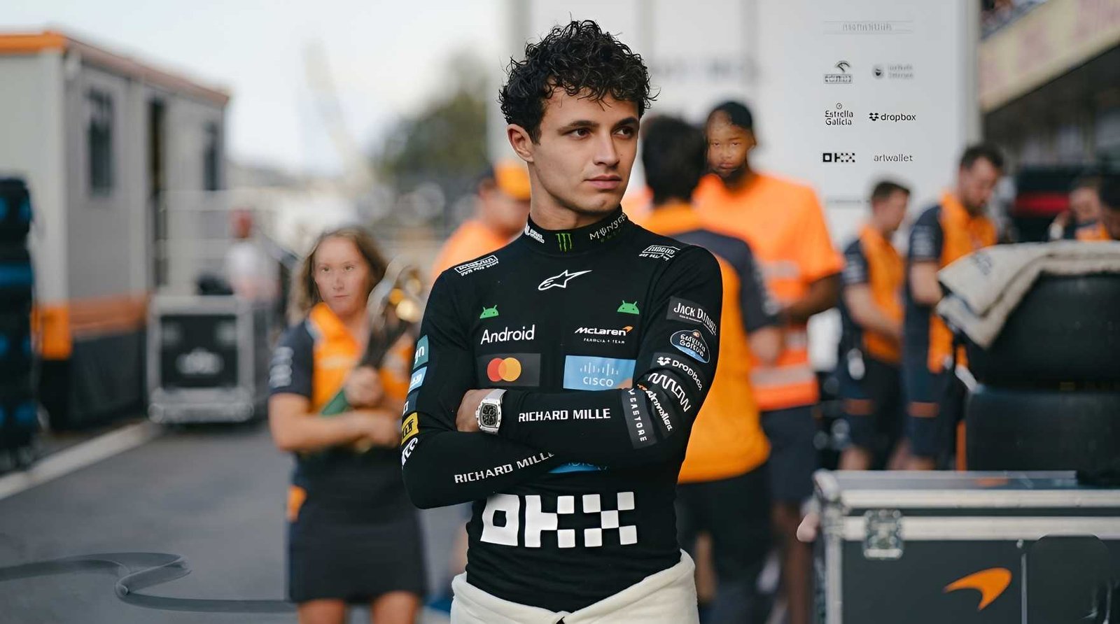 Lando Norris stands with arms crossed wearing McLaren F1 team gear with OKX and Android sponsors in the paddock during the 2026 season