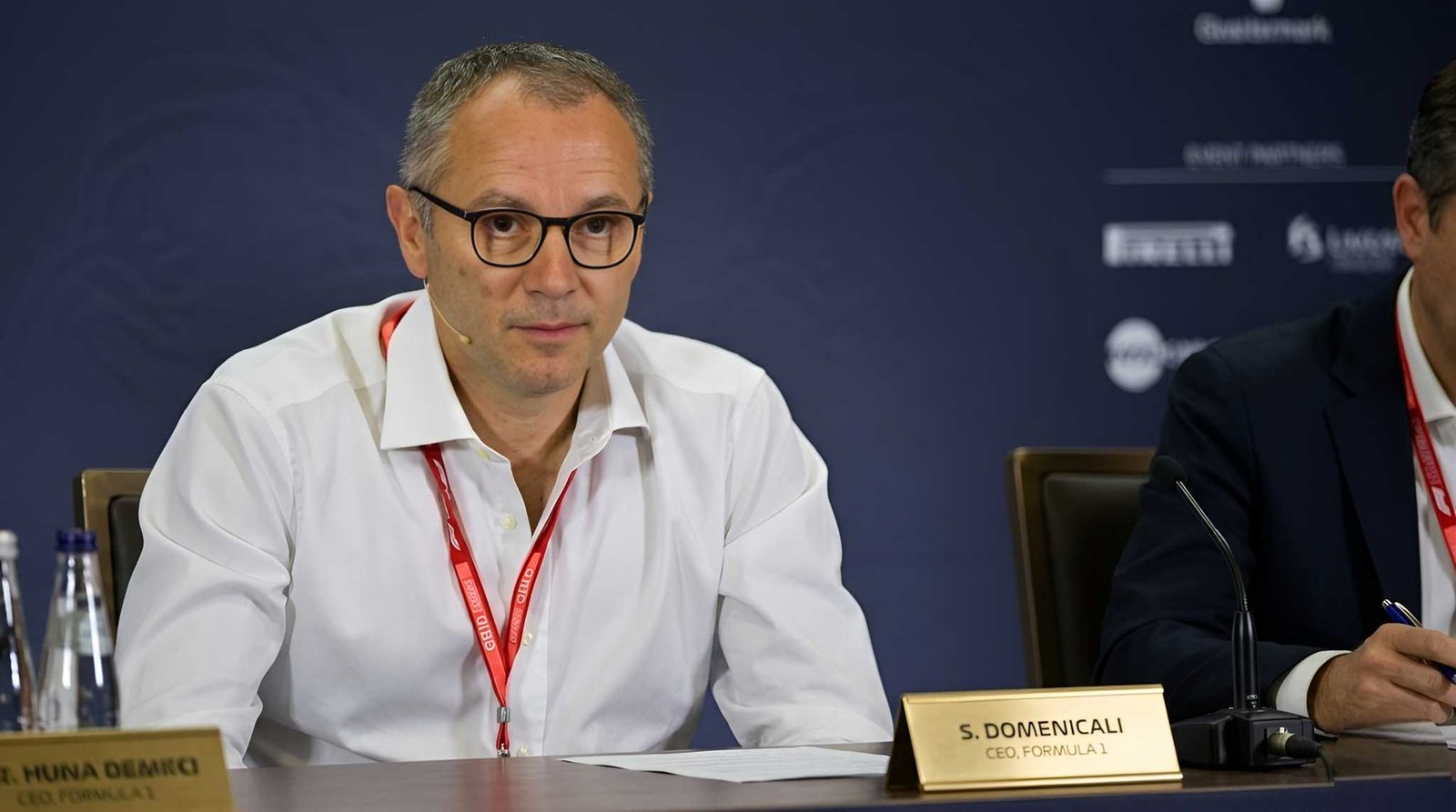 Stefano Domenicali, CEO of Formula 1, seated at a press conference table with nameplate reading S. DOMENICALI, wearing a white shirt and red F1 lanyard